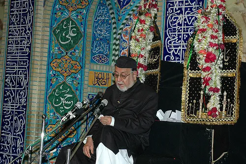 Maulana Mirza Mohammed Athar addressing a majlis at the Mughal Masjid, Mumbai, seated before tiled walls and garlanded alams